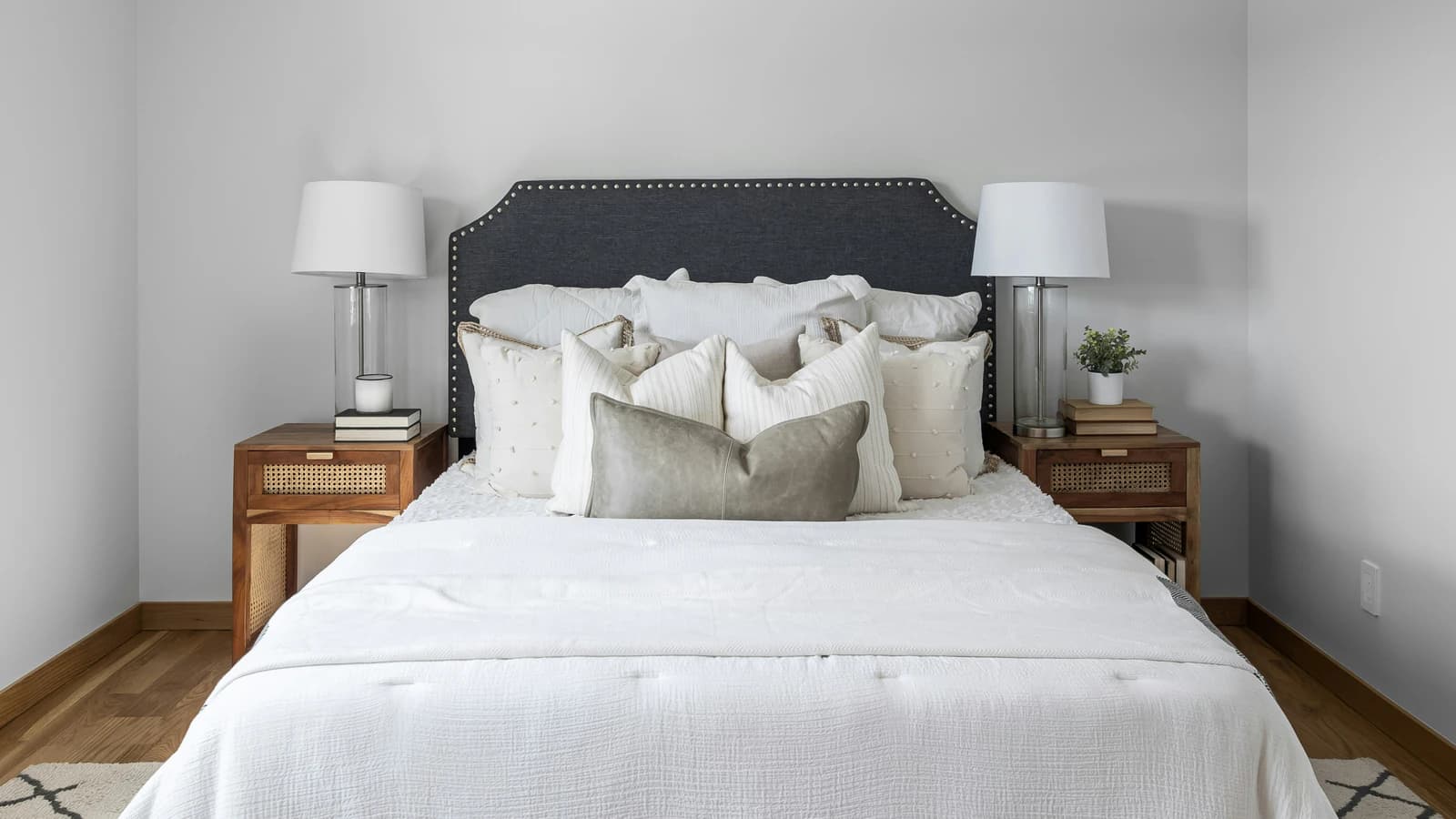 A made bed with a white textured duvet and layered cream and grey pillows, between rattan-front wooden nightstands with white lamps