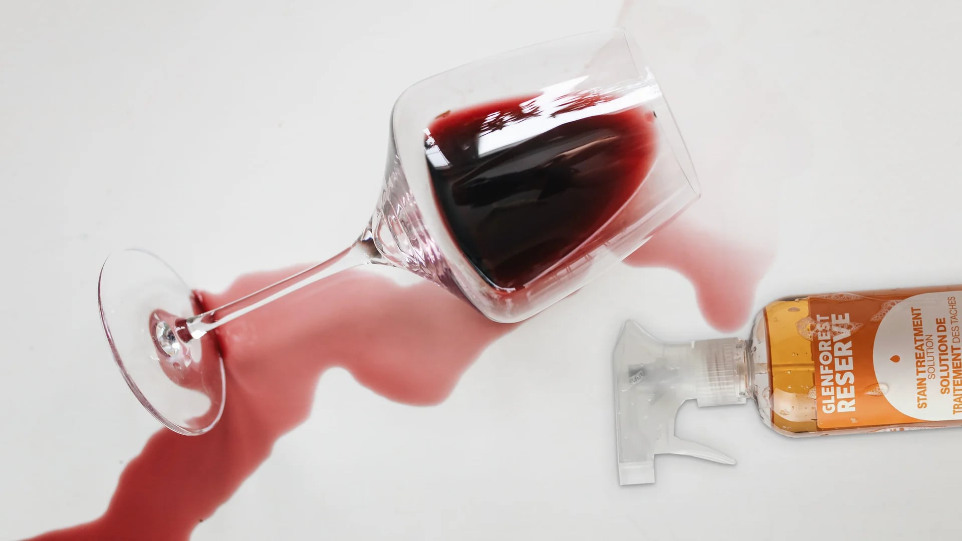 Red wine running from a tipped glass across a white surface, beside a spray bottle of Glenforest Reserve stain treatment solution