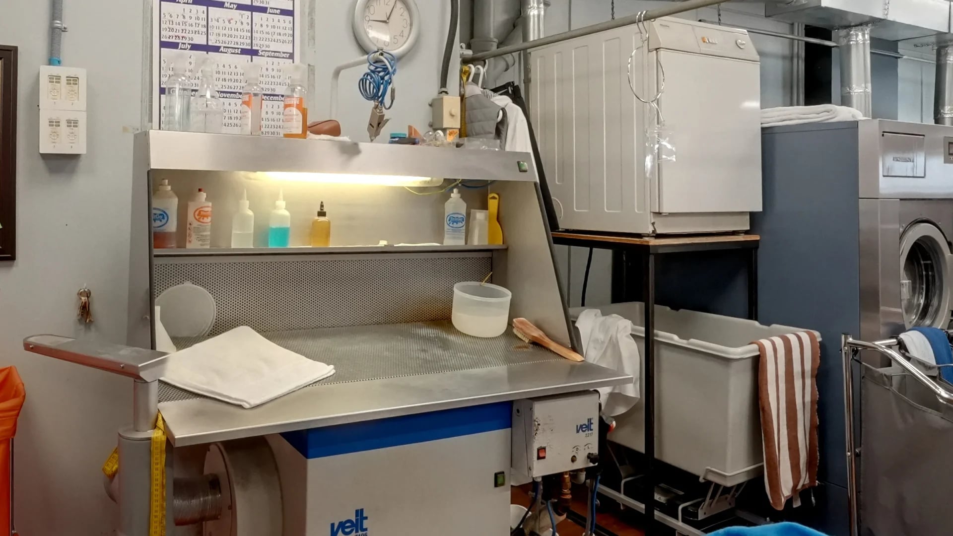 A lit stain-spotting board with spotting bottles on its shelf and a folded towel on its steel table