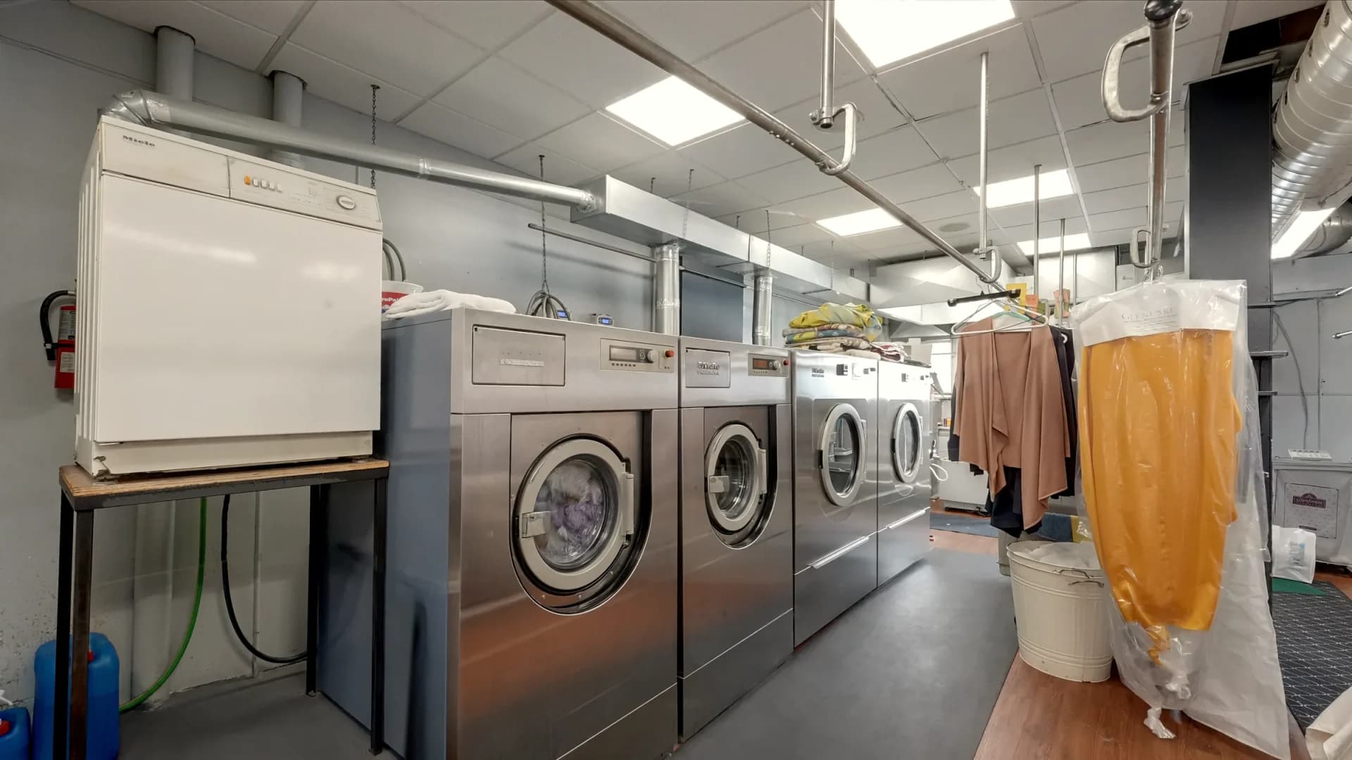 Stainless front-loading washers and a dryer in the Glenforest wet-cleaning room, beside a garment hanging in a clear cover